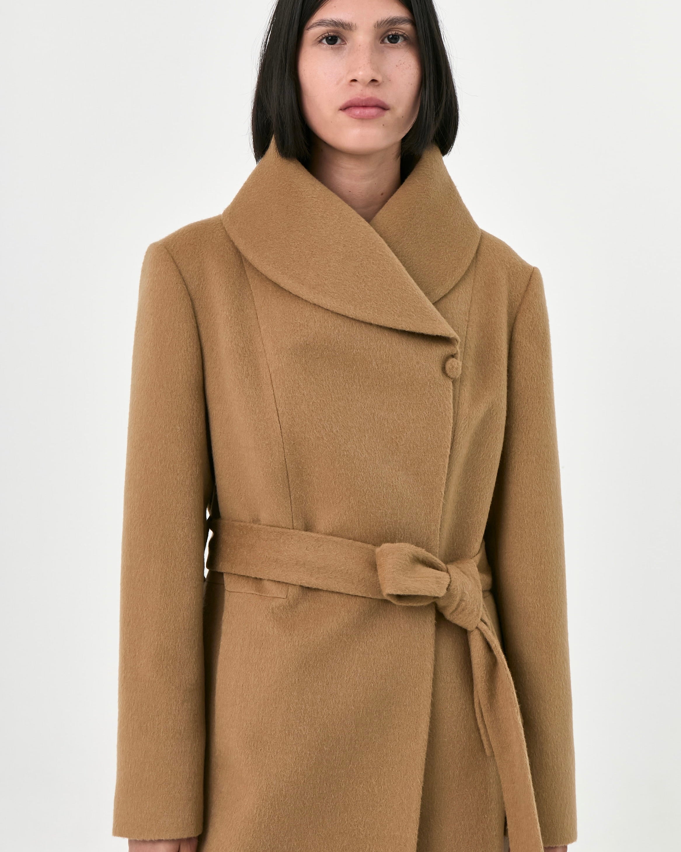 A woman wearing a camel baby alpaca coat with a wrap collar, button closure, and matching waist tie belt.