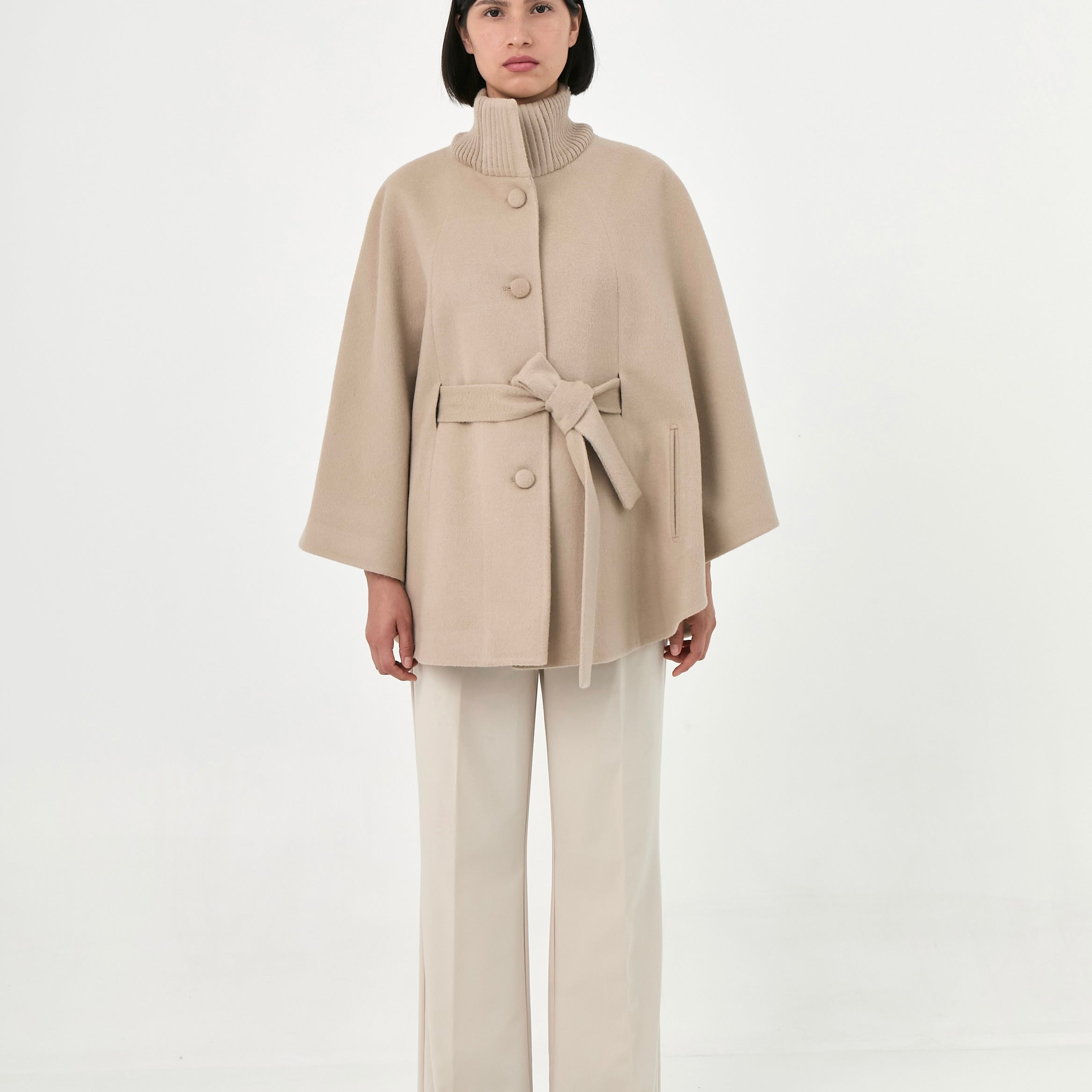 A model wearing a beige baby alpaca cape with a high ribbed collar, a matching belt, buttons front, and side pockets.