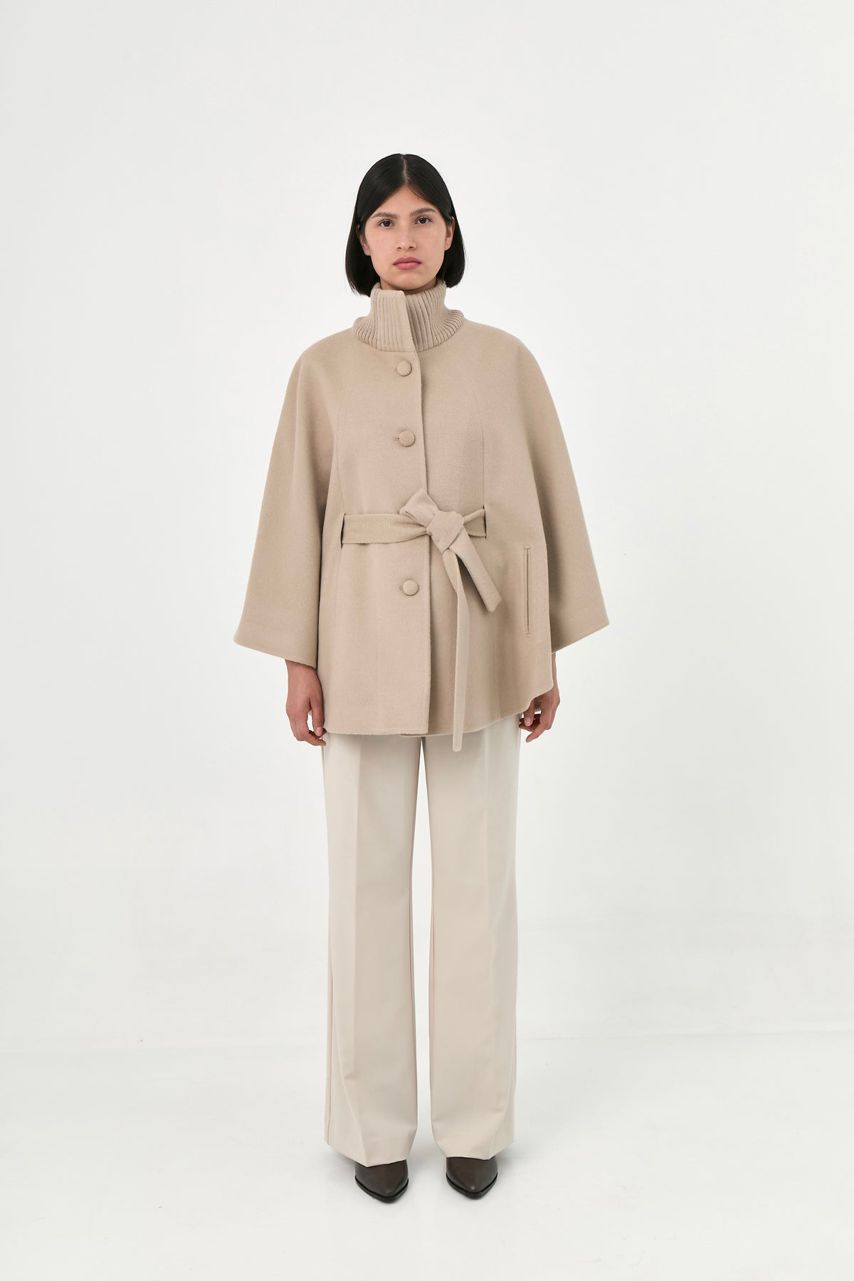 A model wearing a beige baby alpaca cape with a high ribbed collar, a matching belt, buttons front, and side pockets.