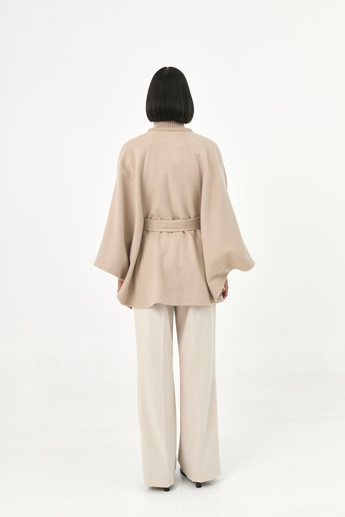 A model viewed from behind wearing a beige baby alpaca cape with a high ribbed collar and a matching belt.