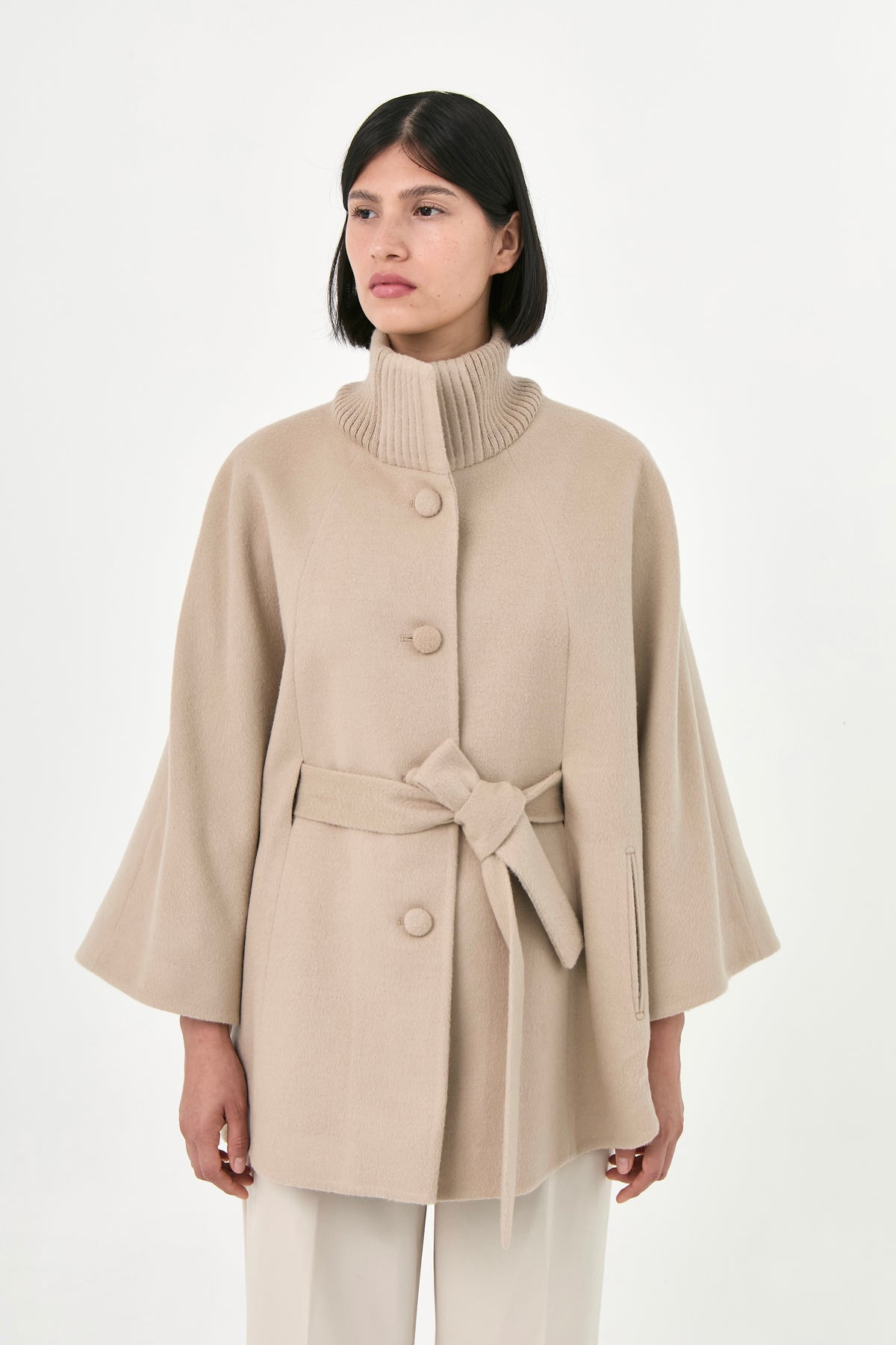 A model wearing a beige baby alpaca cape with a matching belt, buttons front, and side pockets.