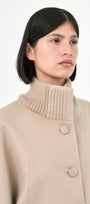 A model wearing a beige baby alpaca cape with a high ribbed stand collar, and buttons front.