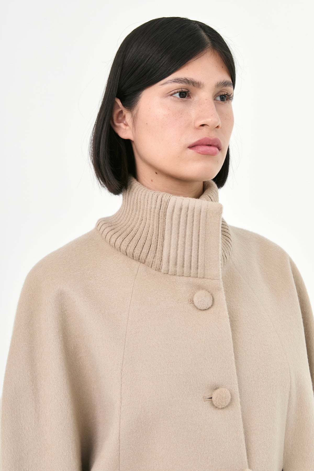 A model wearing a beige baby alpaca cape with a high ribbed stand collar, and buttons front.
