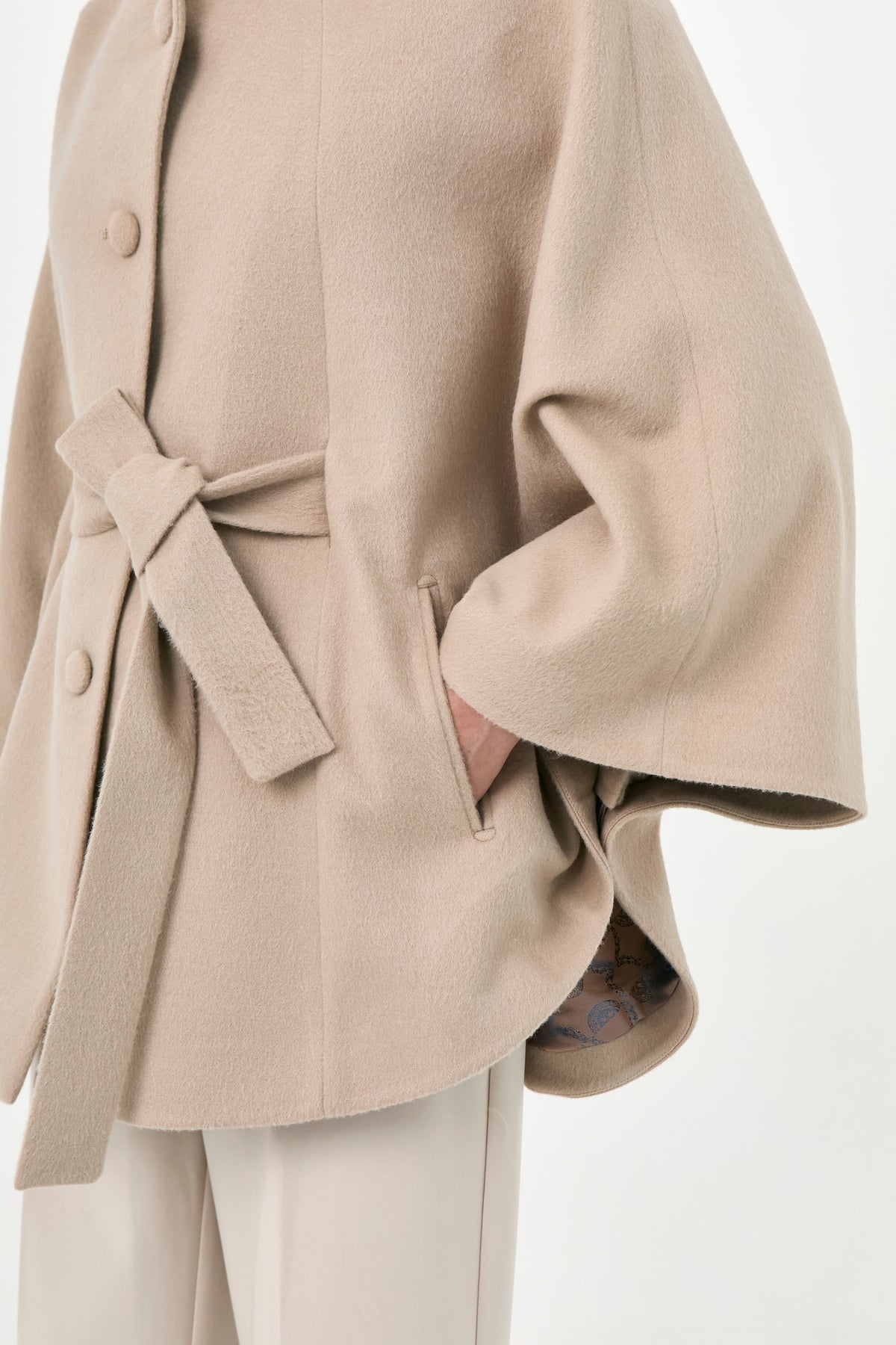 A close-up of a beige baby alpaca cape with a wiast tier, buttons front, and side pockets.
