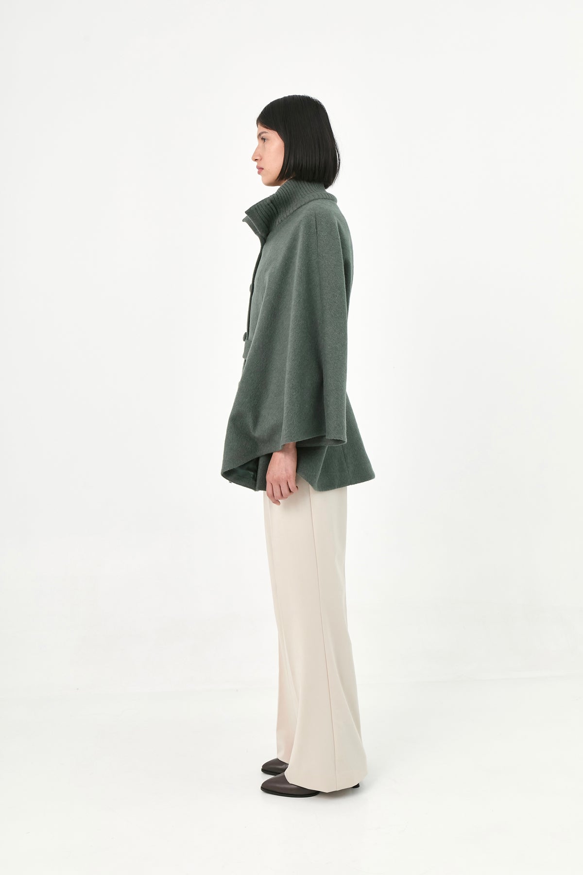 A model shown from the side profile wearing a green baby alpaca cape with a high ribbed stand collar.
