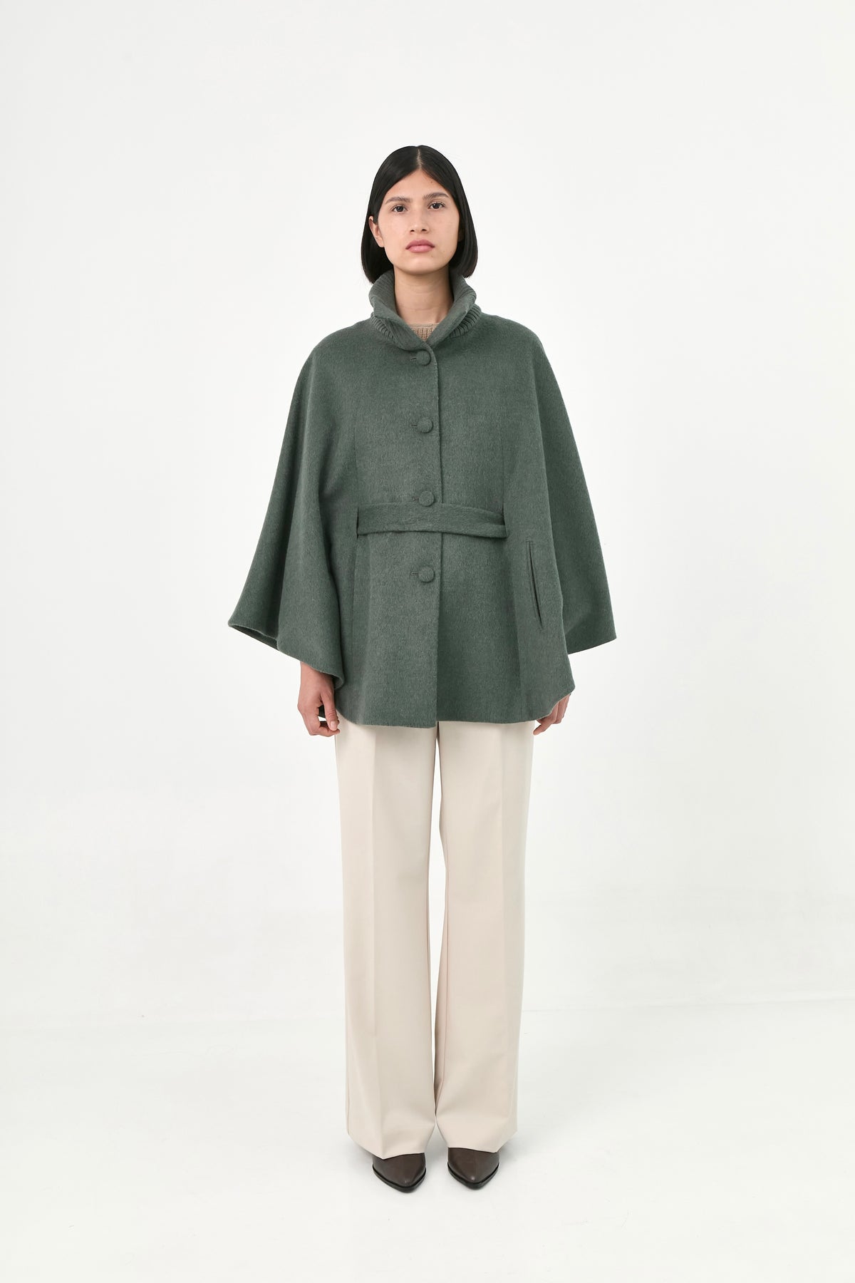 A model wearing a green baby alpaca cape with a high ribbed stand collar, buttons front, and matching waist tie.