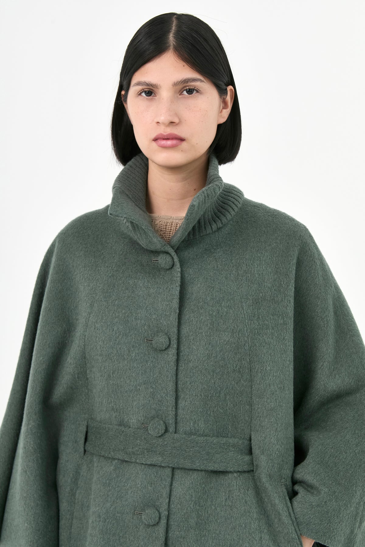 A model wearing a green baby alpaca cape with a high ribbed stand collar, buttons front, and matching waist tie.