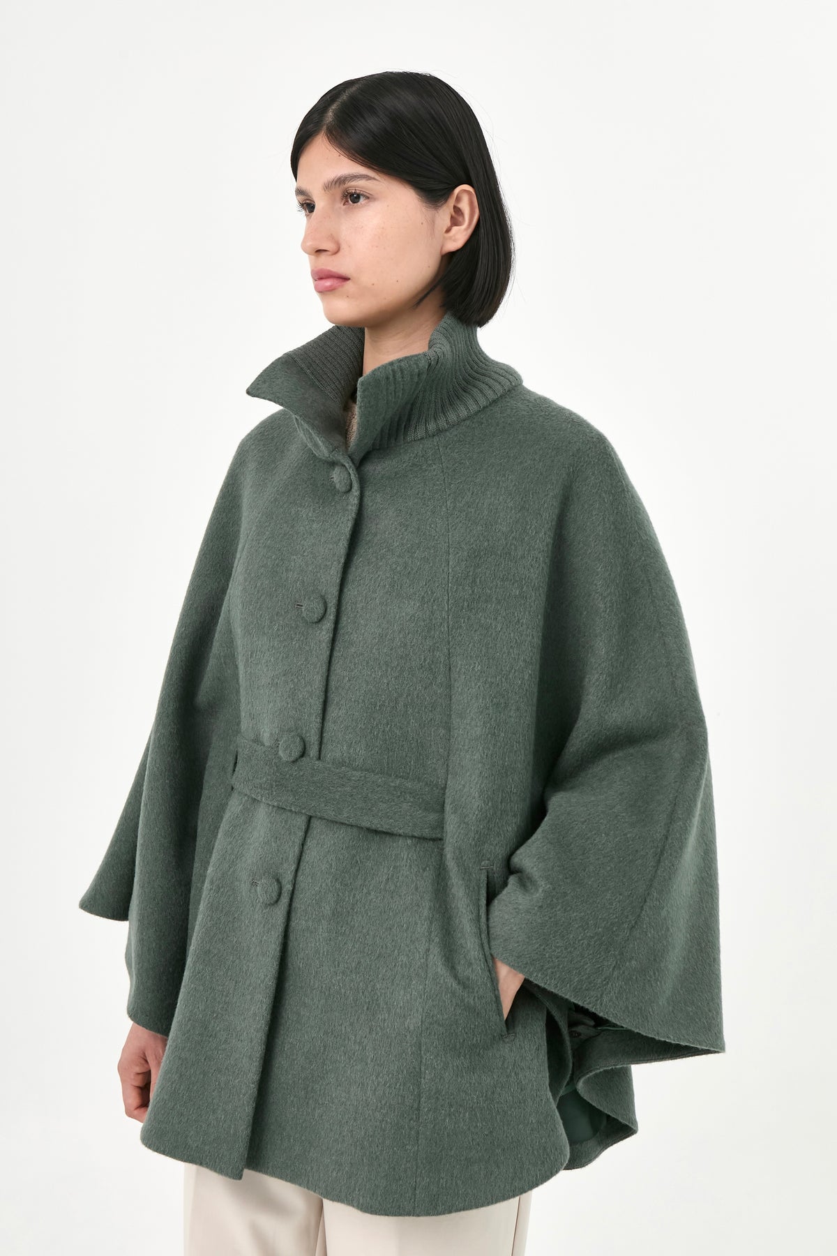 A model wearing a green baby alpaca cape with a high ribbed stand collar, buttons front, and matching waist tie.
