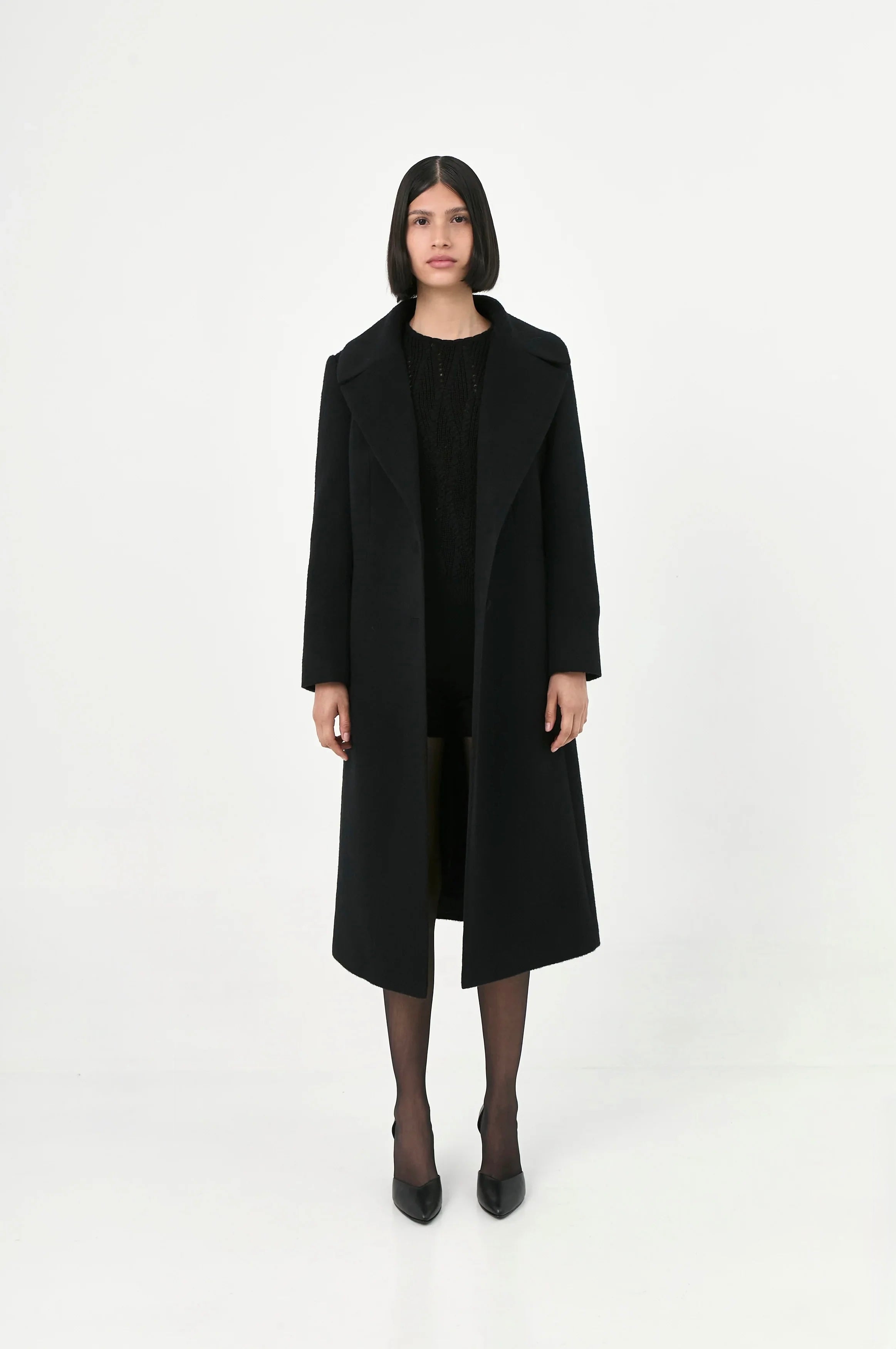 A model wearing a long black baby alpaca coat with a wide pointed lapel.