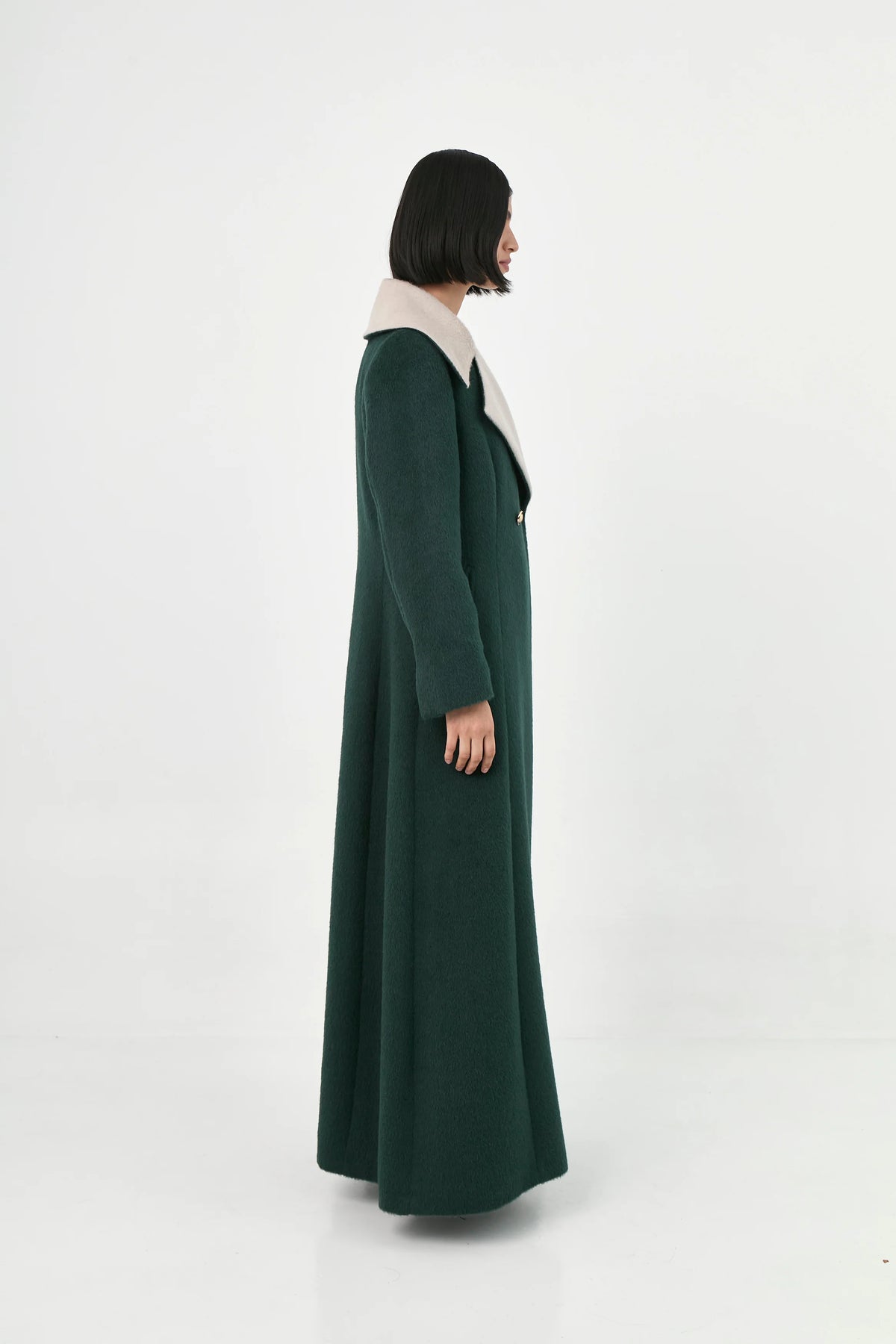 A model shown from the side profile wearing a long, dark green suri alpaca coat with a contrasting light beige collar.