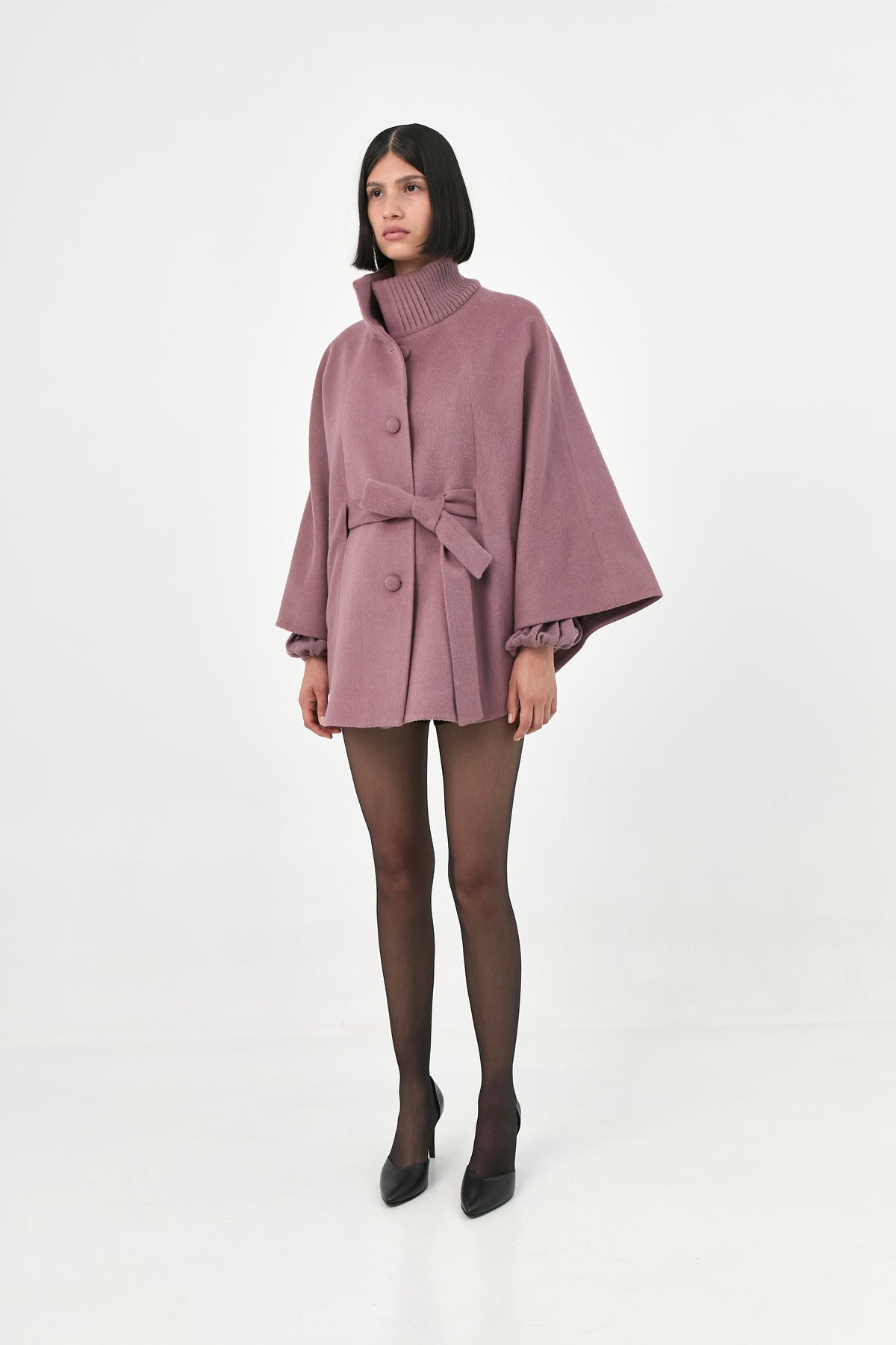 A model wearing a mauve baby alpaca cape with a high ribbed collar and a belted waist.