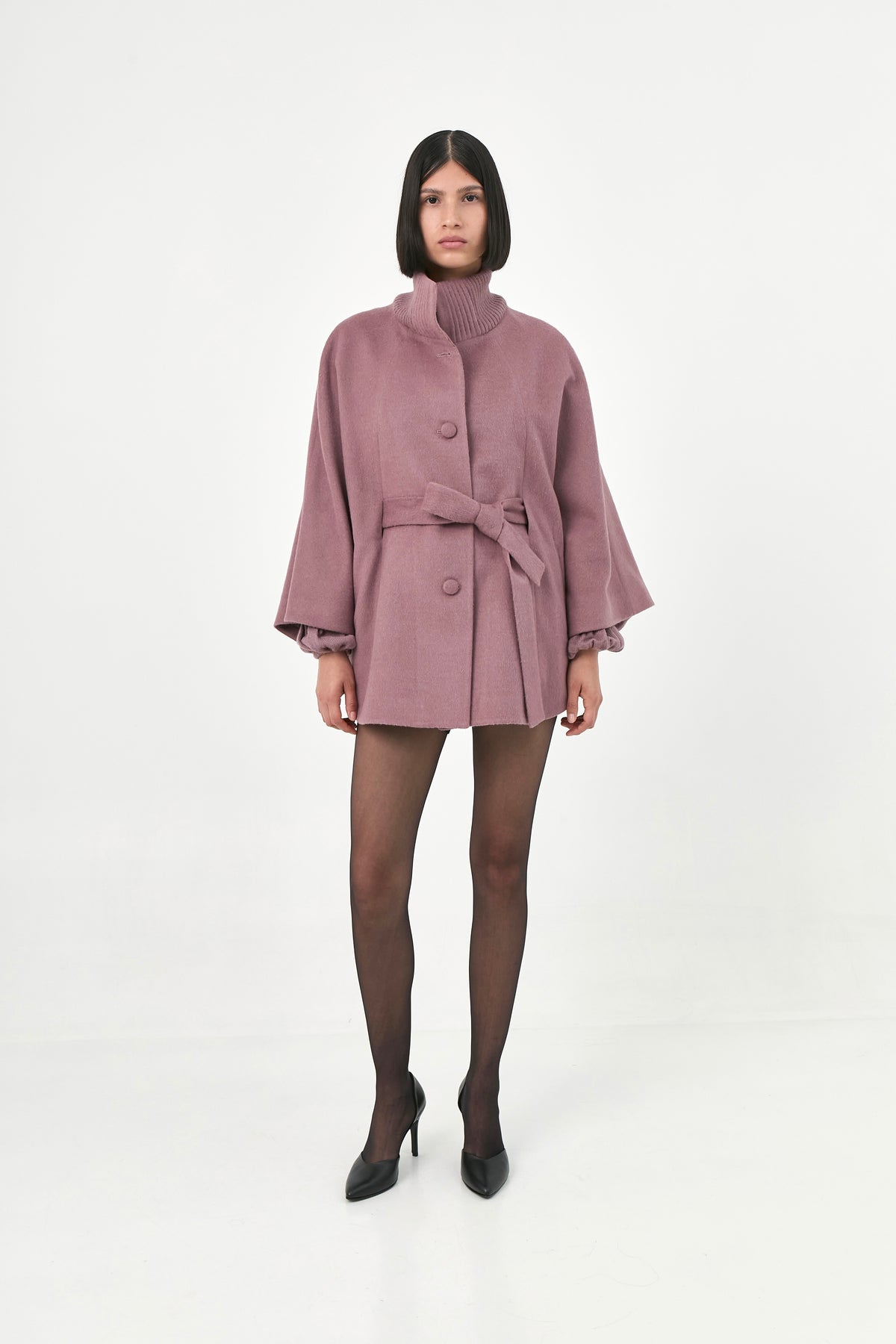 A model wearing a mauve baby alpaca cape with a high ribbed turtleneck collar and a belted waist.