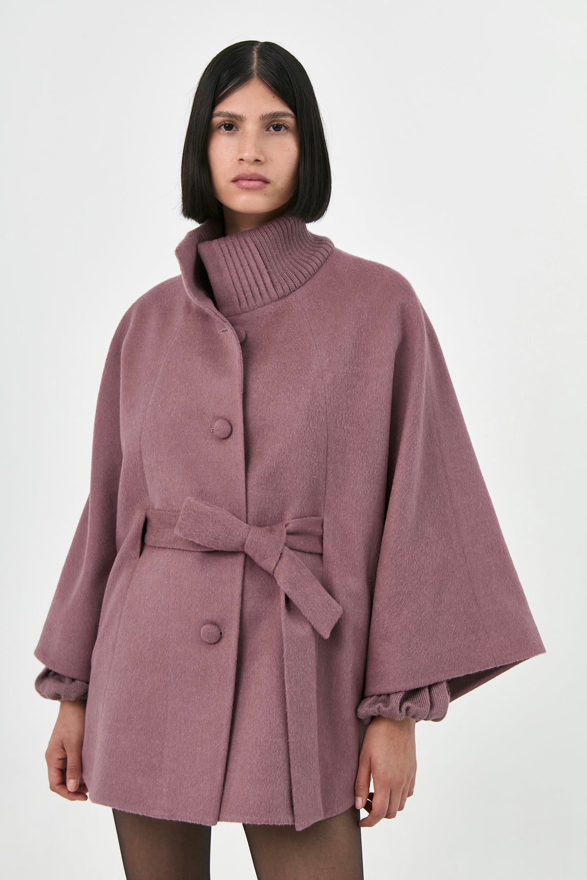 A model wearing a rose mauve baby alpaca cape with a turtleneck ribbed collar and a belt.