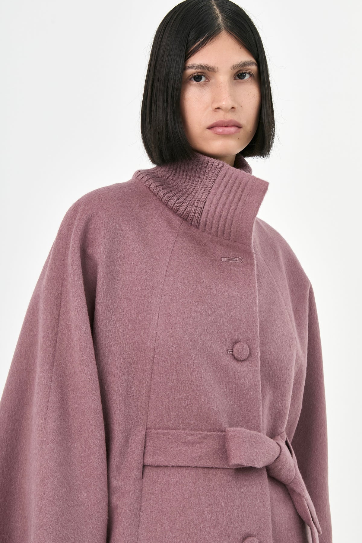 A model wearing a mauve baby alpaca cape with a high ribbed turtleneck collar, and a belted waist.