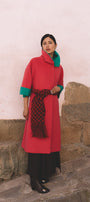 Woman wearing a red coat with green trim and a patterned scarf, standing against a stone wall in Cusco.