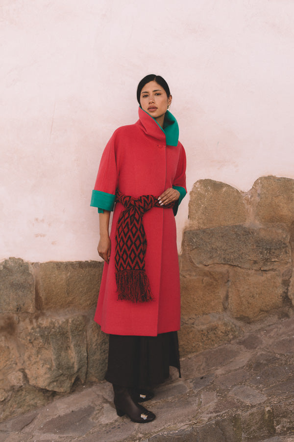 Woman wearing a red coat with green trim and a patterned scarf, standing against a stone wall in Cusco.