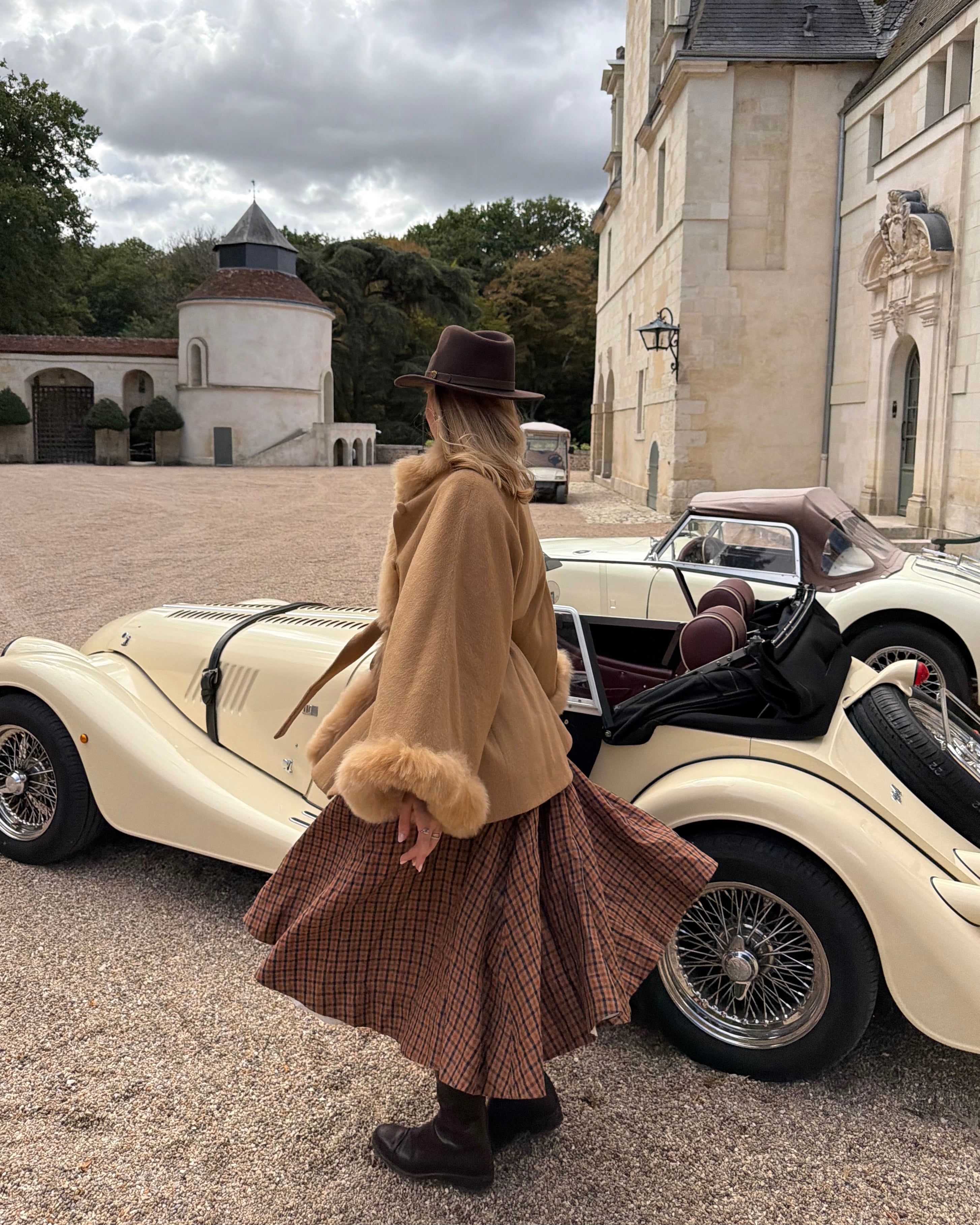 A woman wearing a baby alpaca cape with fur , a brown plaid skirt, and a brown hat stands next to a cream vintage convertible car in front of a large stone building.